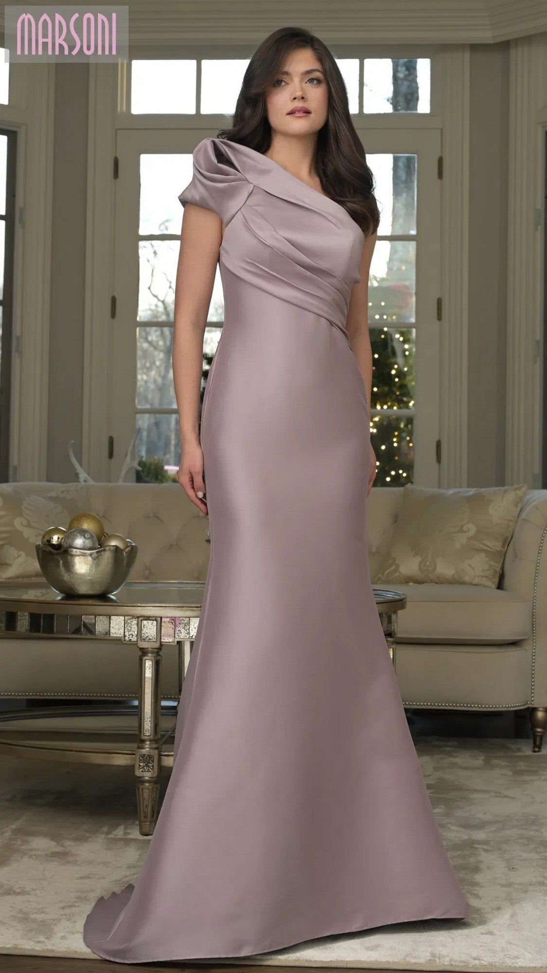 A woman poses indoors in a formal living room with neutral furniture, wearing the Marsoni MV1358 long formal dress in dusty rose, featuring a floor-length silhouette, one-shoulder design, and elegant draped detailing.